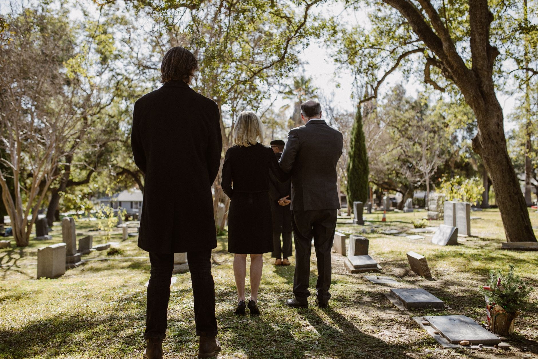 A family of three stands with backs to the camera in a sunlit cemetery, facing a graveside service.